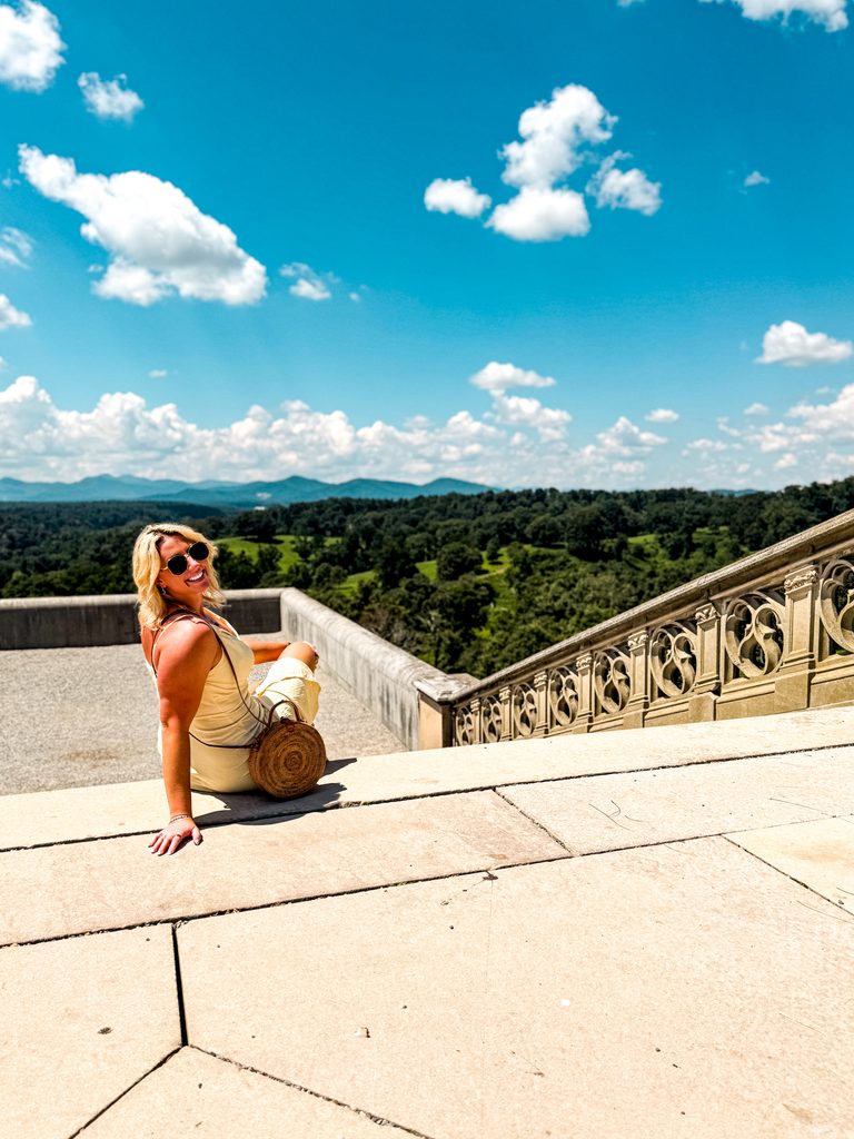 Amanda Dante sitting on stone steps beside an ornate railing, overlooking green hills and distant mountains beneath a blue sky.