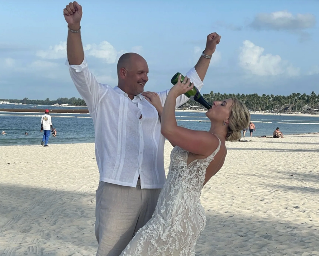 Bride drinking from a green bottle beside a groom raising his arms on a sandy beach, with the ocean and palm trees behind them.