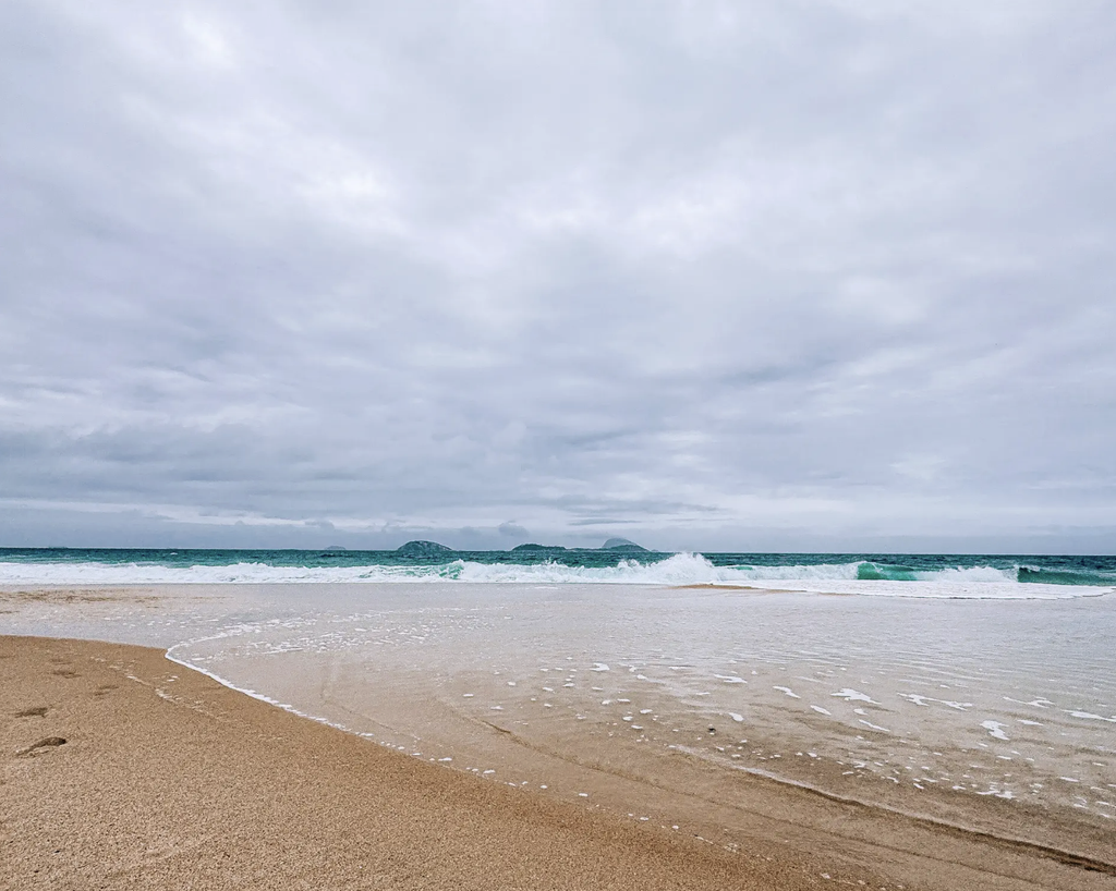 Waves washing onto a sandy beach in Rio de Janeiro, with offshore islands beneath a cloudy sky.