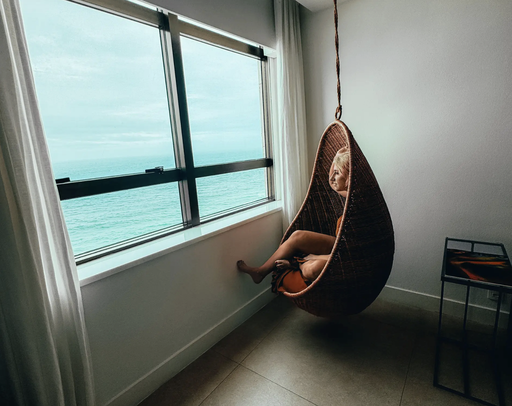 Amanda Dante sitting in a hanging wicker chair beside an ocean-view window in Rio de Janeiro.