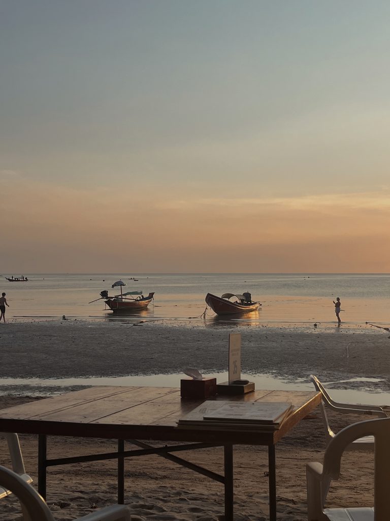 Wooden long-tail boats float near a sandy shoreline beneath a peach-colored evening sky in Thailand.