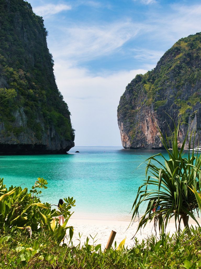 Turquoise water between towering limestone cliffs, with a sandy beach and tropical plants in the foreground.